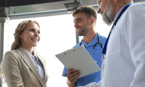 A doctor and a nurse with a clipboard speak to a woman in business clothes.
