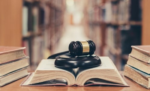 A gavel rests on an open book in the middle of a table with stacks of books on either side, with a library row stretching behind it.