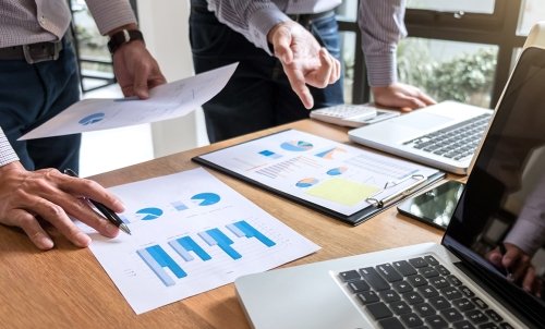 Two people stand at a desk with laptops and papers displaying bar graphs and pie charts, discussing.