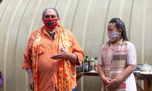 Two native people give a presentation in front of a green house and a table with cooking ingredients on it.