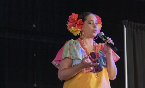 A woman in colorful costume and flowers in her hair speaks or sings into a microphone