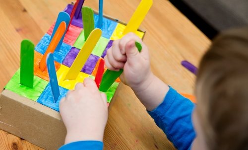 A child plays with a brightly colored toy: a box with colored squares and matching colored popsicle sticks in each.