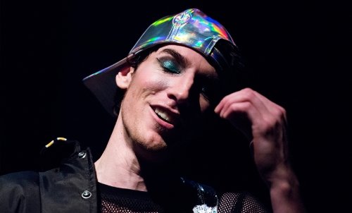 Close up of an actor, whose head is titled, hand raised, and eyes closed.  He has teal eyeshadow, a brightly colored baseball cap worn backwards, and a smirk.