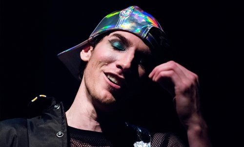 AcademicProgram_Theatre.jpg Close up of an actor, whose head is titled, hand raised, and eyes closed. He has teal eyeshadow, a brightly colored baseball cap worn backwards, and a smirk.