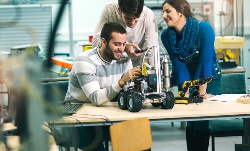 Three students work on a small machine on wheels on a desk in a lab.