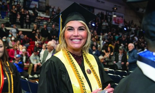 A graduate in cap and gown smiles during the ceremony with a court full of people in bleachers behind her.
