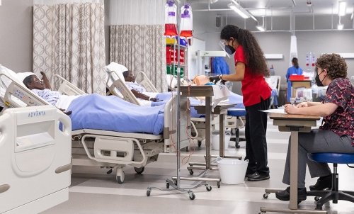 A student works at the bedside of a dummy patient in the nursing lab while an evaluator looks on.