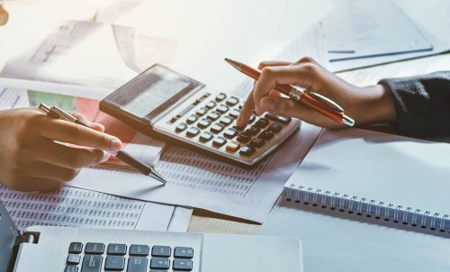 Close up of a hand holding a pencil working on a calculator with a laptop, lots, of paper, and another hand with a pen around it.
