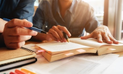 Close up of students working on a bookmarked book, with pens in hand.
