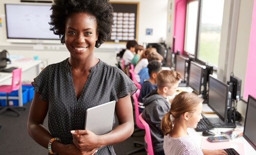 A teacher smiles in the foreground while children work on computers in class in the background.