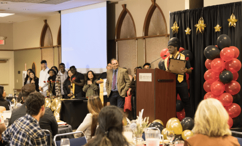 A student stands on a decorated stage to speak to a seated audience, with a line of his peers to his side