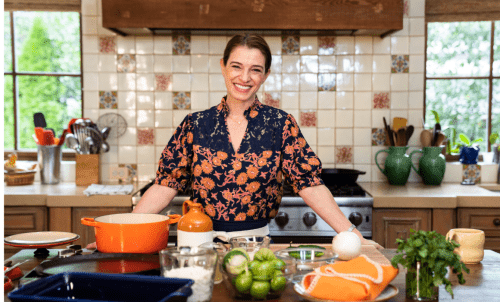 Pati Jinich stands at a counter laden with cooking implements, with her kitchen windows behind