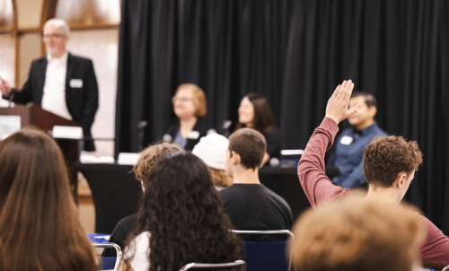 A in a seated crowd raises their hand, with a panel on stage