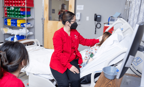 A student in red scrubs and a mask sits on a hospital bed with a training dummy in a nursing lab classroom