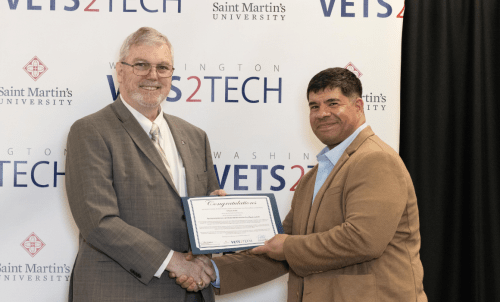 A graduate shakes hands with a dean holding a certificate, the WaV2T backdrop behind