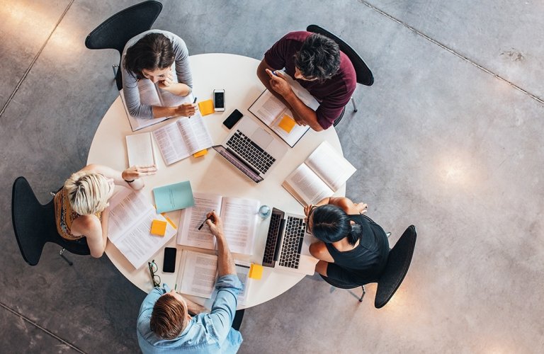 Overhead view of five students studying with books and laptops at a round table.