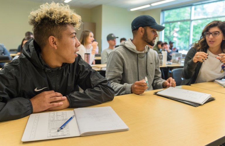 Three students seated in a row at a desk discuss with papers in front of them and a classroom full of other students behind.