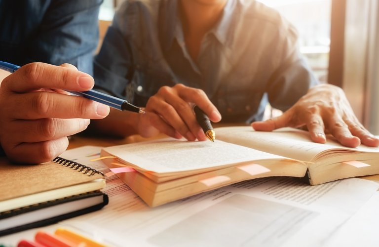 Close up of students working on a bookmarked book, with pens in hand.