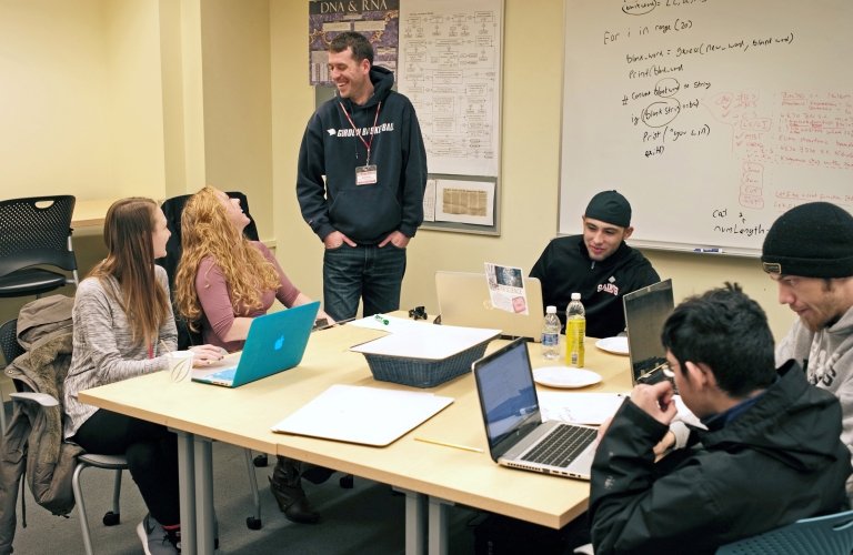 Five students work at laptops on a square table while an instructor smiles in front of a white board.