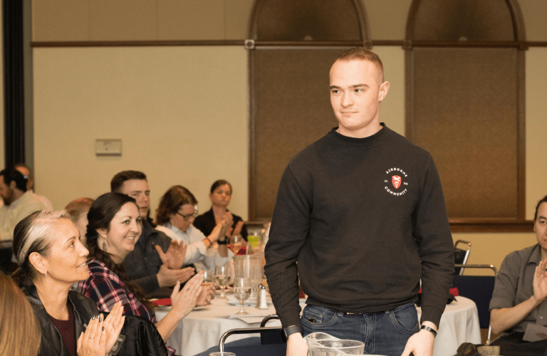 A veteran stands up from a table while a seated audience applauds him.
