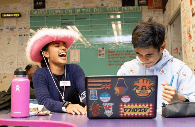 A student in a pink hat laughs with a student in a flowery hoodie in front of a laptop