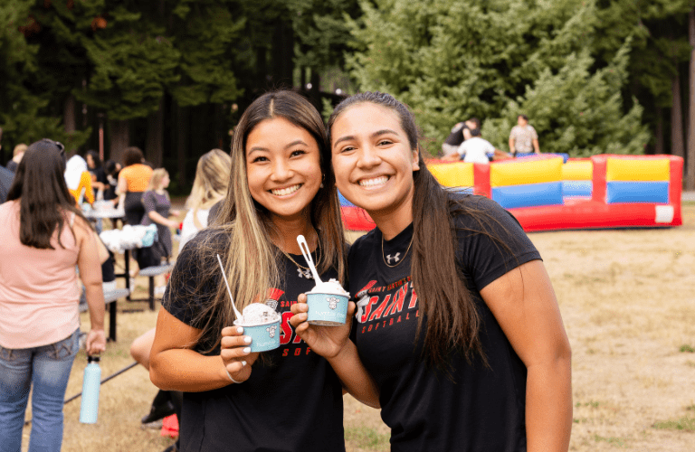 Two students smile with ice cream at an event in front of trees and an inflatable structure