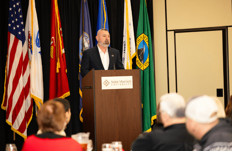 Man stands at podium with American and military flags behind