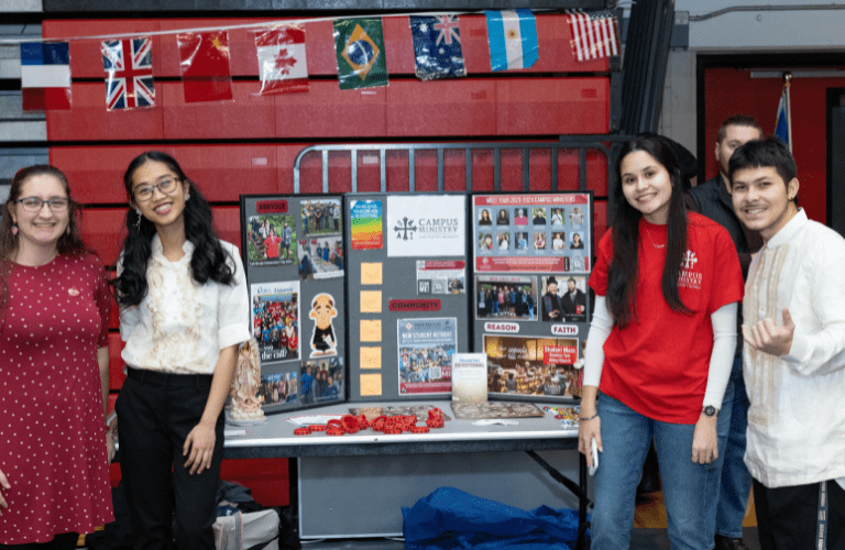 Students in front of Campus Ministry board, many different national flags on bleachers behind