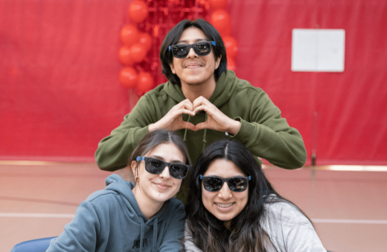 Three students in sunglasses pose in a triangular shape, one making a heart with their hands