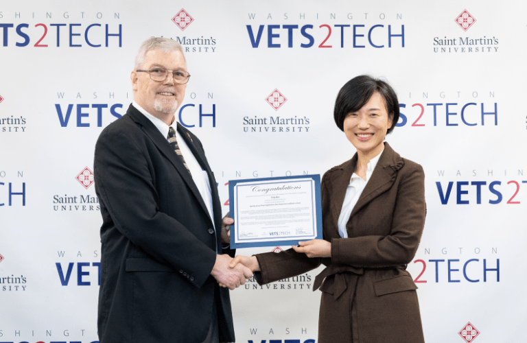 A dean gives a student their diploma in front of a Vets2Tech backdrop