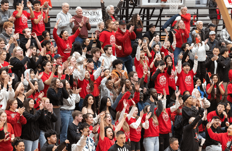 A crowd of students in red shirts cheers from the bleachers at athletics game