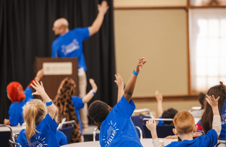Children seated at tables raise their hands as a teacher leads in front