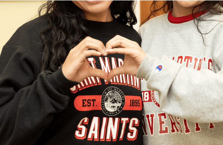 Two students make a heart with their hands in front of Saint Martin's sweatshirts
