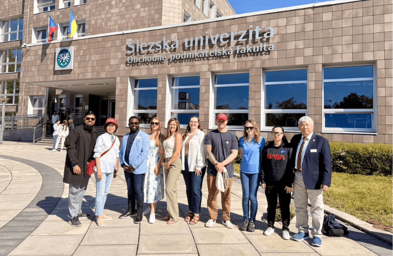A group of students and professors stand in front of a university with name in another language