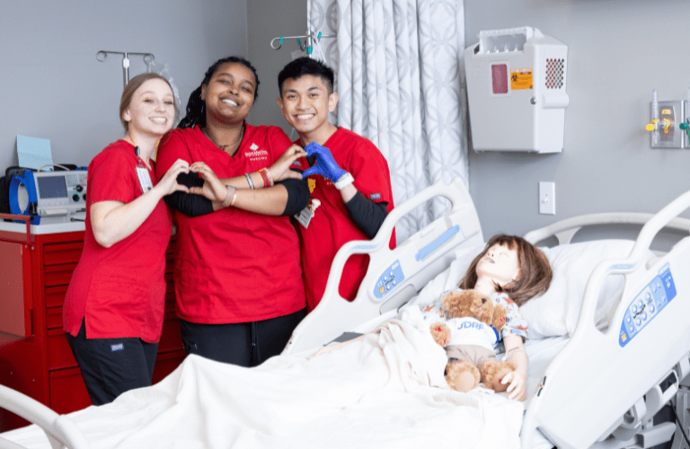 Nursing students in scrubs pose in lab by hospital bed