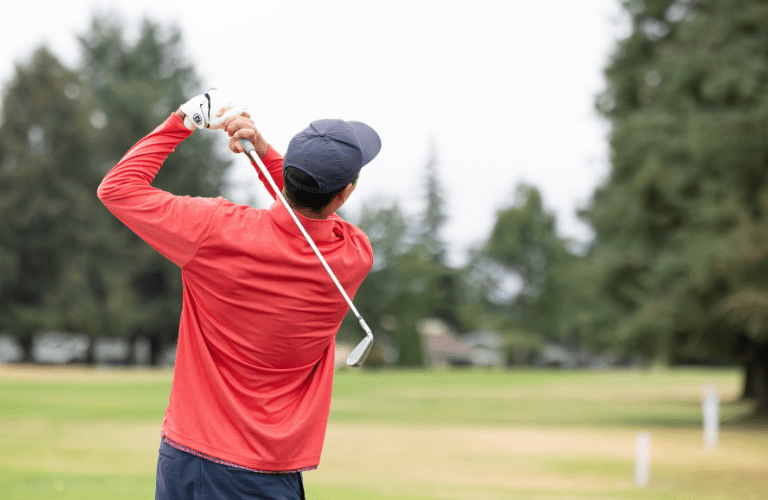 A person wearing red swings a golf club on a tree-lined course