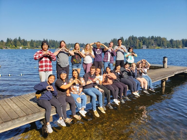 Students at the New Student Retreat sit and stand on a dock stretching out into the lake, making hearts with their hands on a sunny day