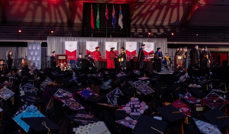 A graduate crosses the commencement stage to receive their degree from the president, decorated graduation caps in the foreground