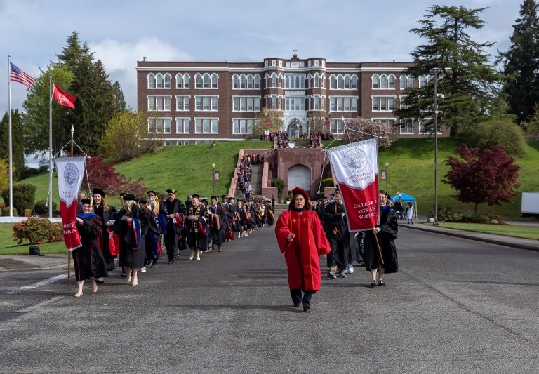 Dr. Tam Dinh leads the line of graduate marching to commencement, graduates in regalia and holding college banners, with Old Main at the top of the grand staircase in the background