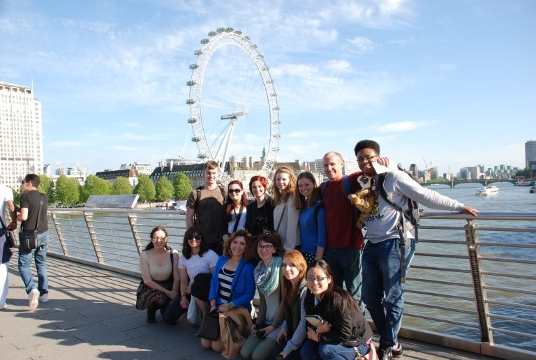 Students pose against the guardrails over a river, with the London Eye in the background