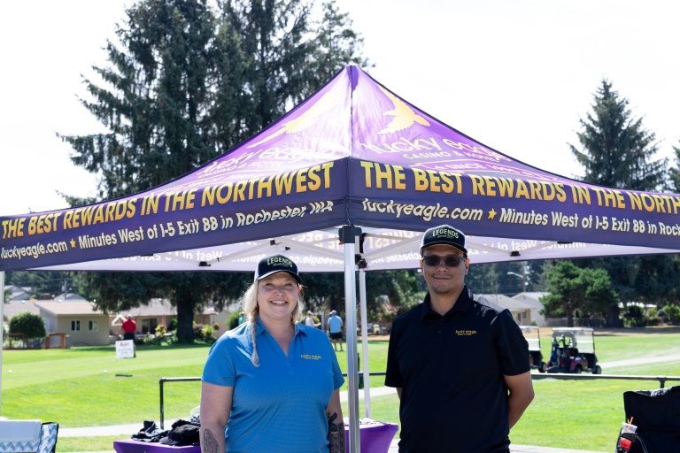 Two people stand below a purple Lucky Eagle awning with the golf course in the background