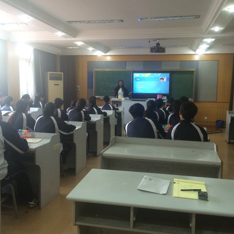 Students in uniforms sit at tables facing the front of a classroom, where a teacher presents