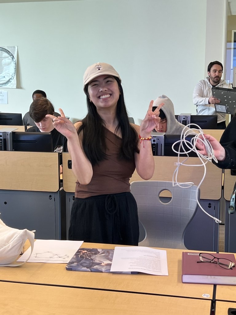 A student standing in a row of desks poses for the camera, her peers working around her