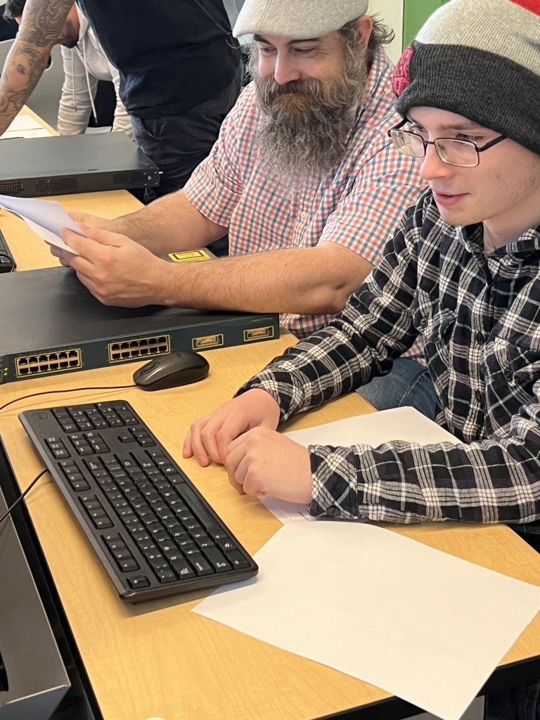 A student works at a computer, with a professor reviewing at side