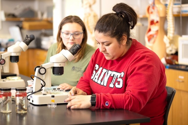 Two students sit at a desk working with microscopes in a lab