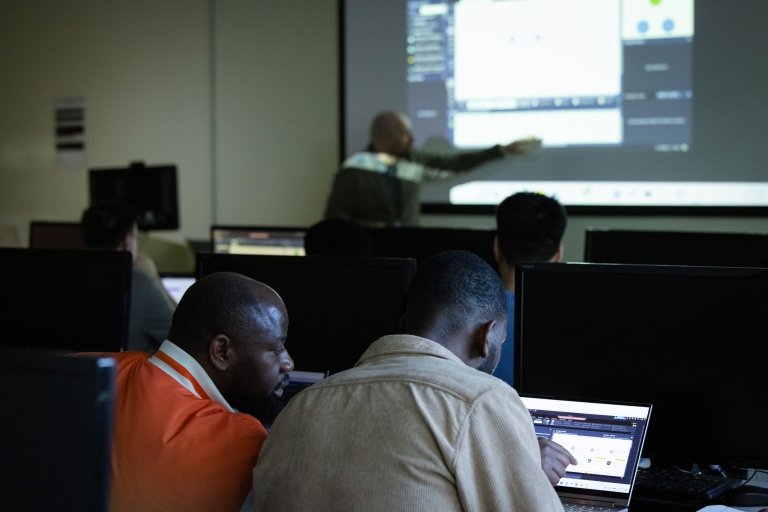 Students look at computers while a professor demonstrates at the front of the classroom