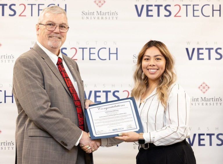 A student shakes hands with professor while holding degree in front of WA Vets 2 Tech background