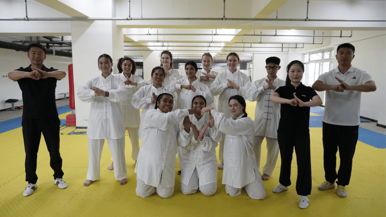 Students in white robes do hand salute with tai chi teachers in a room