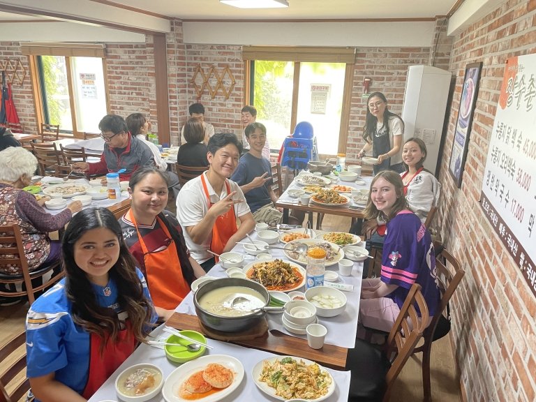 Students in aprons sit at tables with lots of food in a dining room