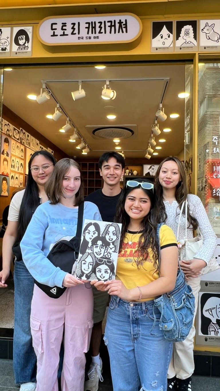 Students pose with a caricature drawn of them in front of a building with caricatures on the wall